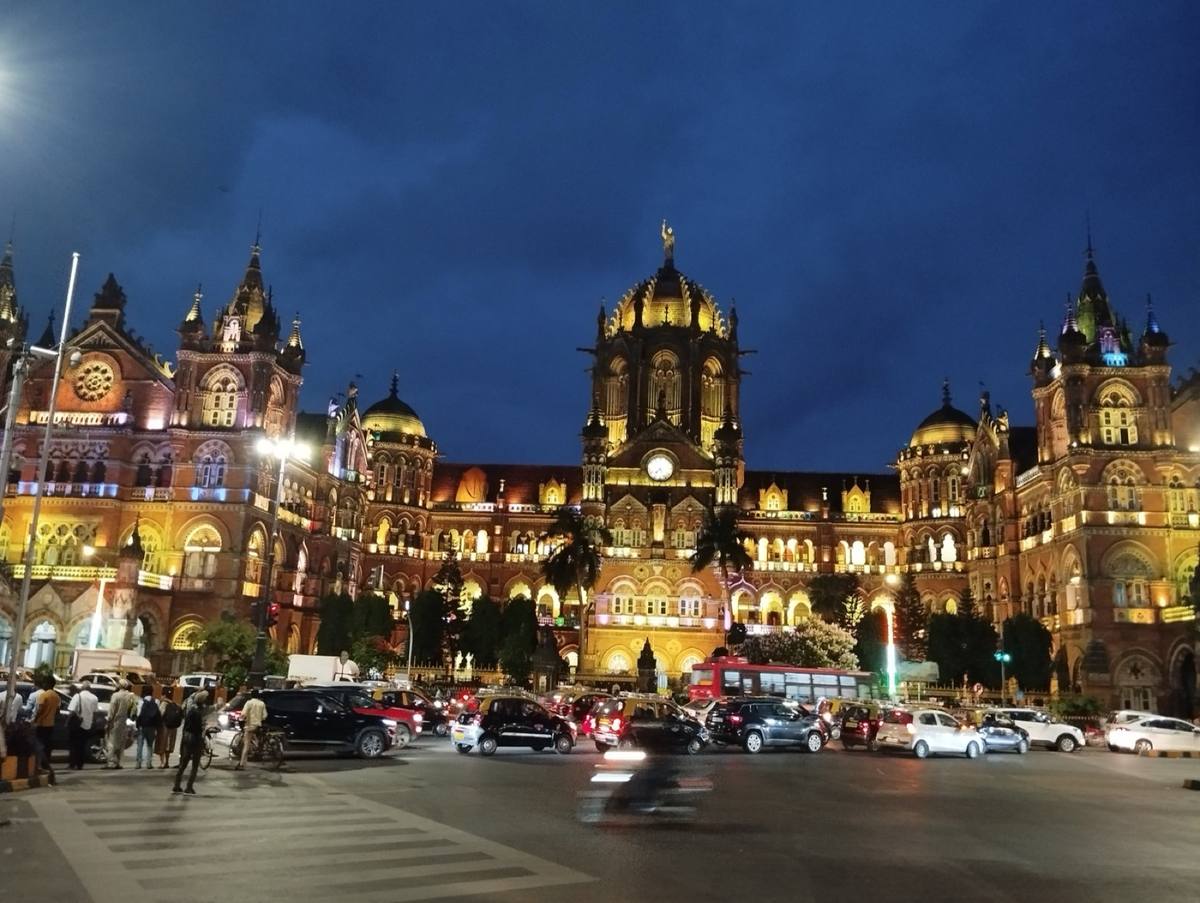 Colour photograph of a Gothic Revival railway station with domes and spires floodlit gold under a blue evening sky, traffic in front