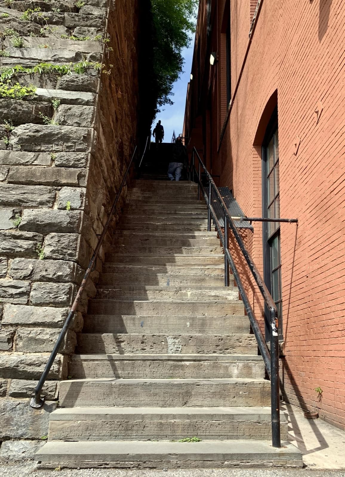 A long, narrow flight of stone steps rising between a rough stone wall and a red-brick wall, a lone figure at the top