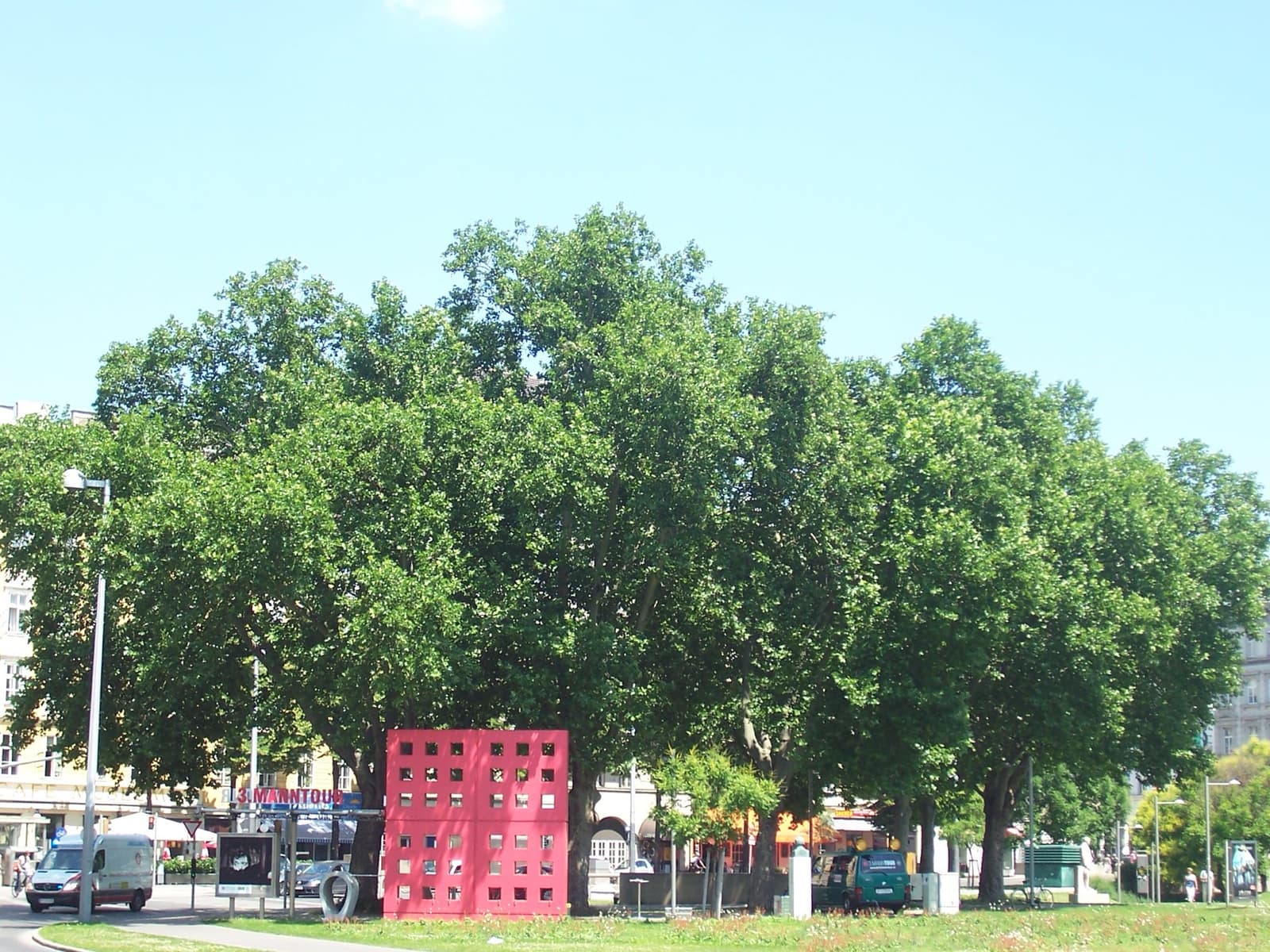 A park of tall trees with a red metal grate structure on the lawn