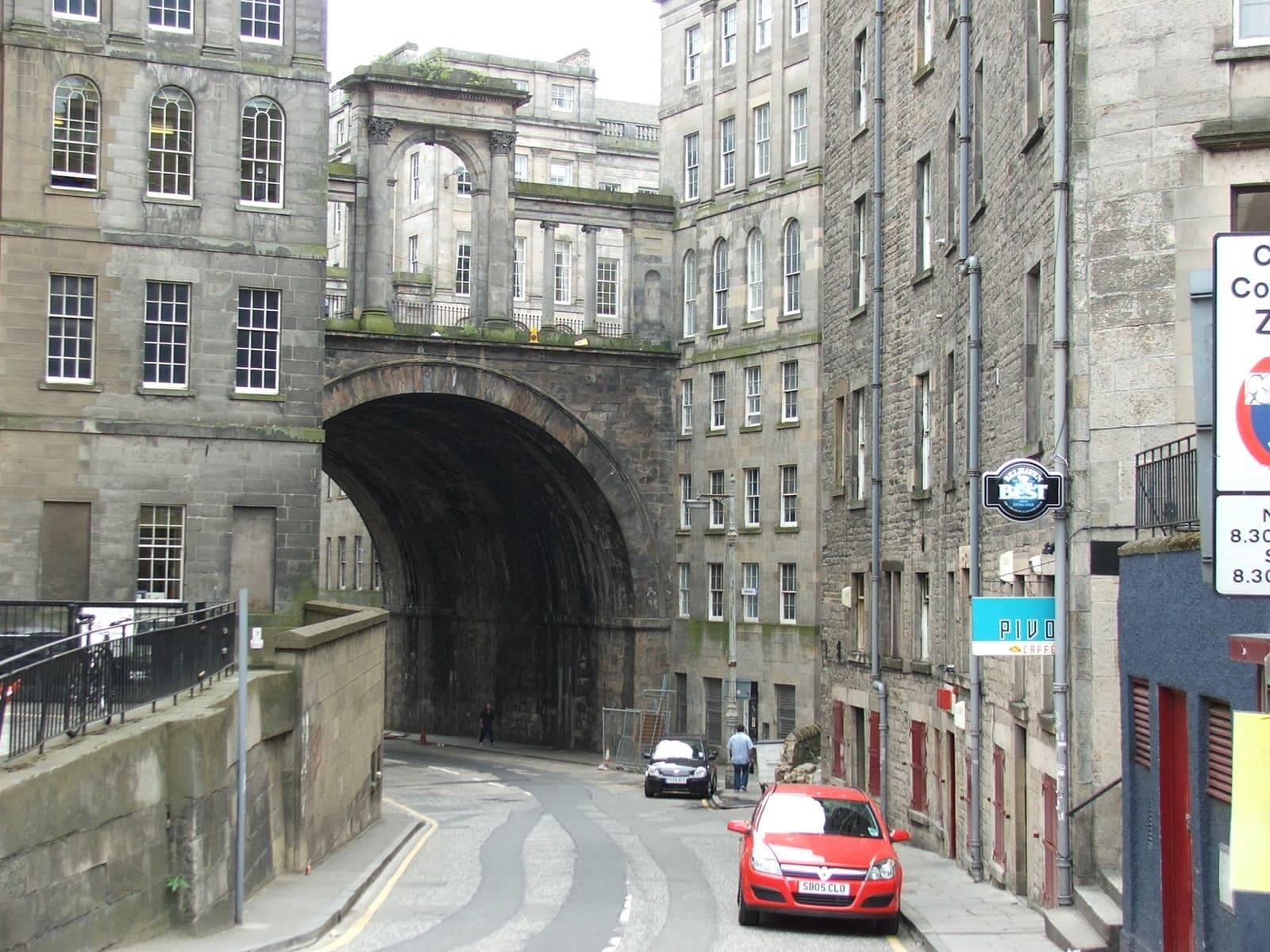 A tall stone arch carrying a road over a narrow sloping street, a bar sign on the wall
