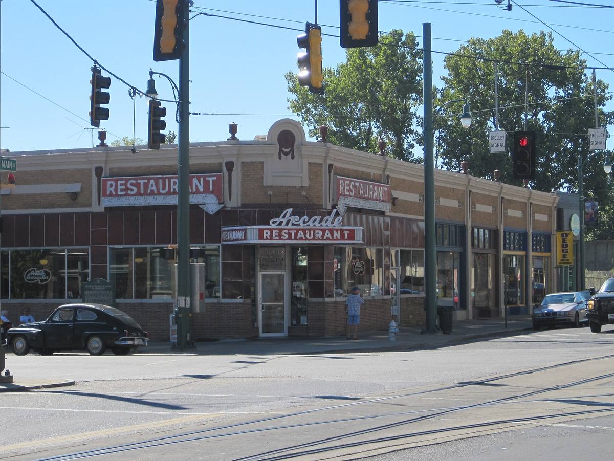 A low corner diner with a neon Restaurant sign and an old black car parked outside