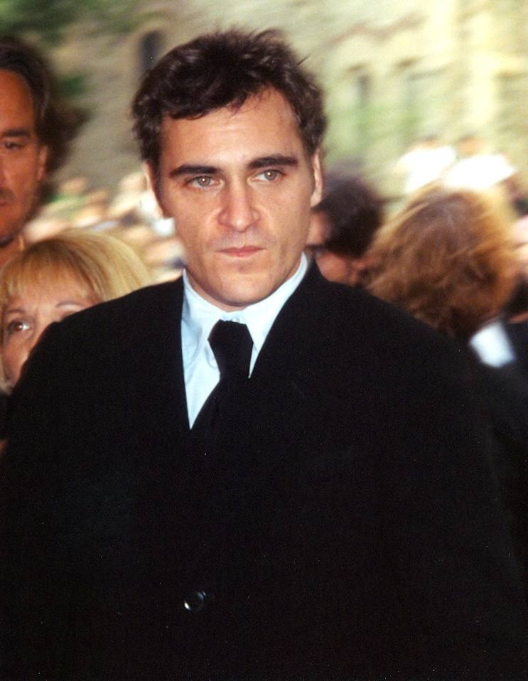 A dark-haired man in a black suit and tie looks into the camera, a crowd blurred behind him