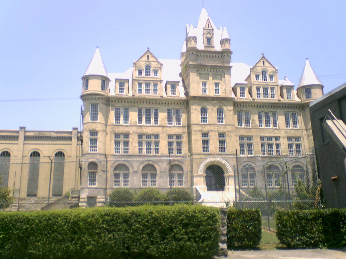 A turreted stone castle-like prison building behind a hedge and fence