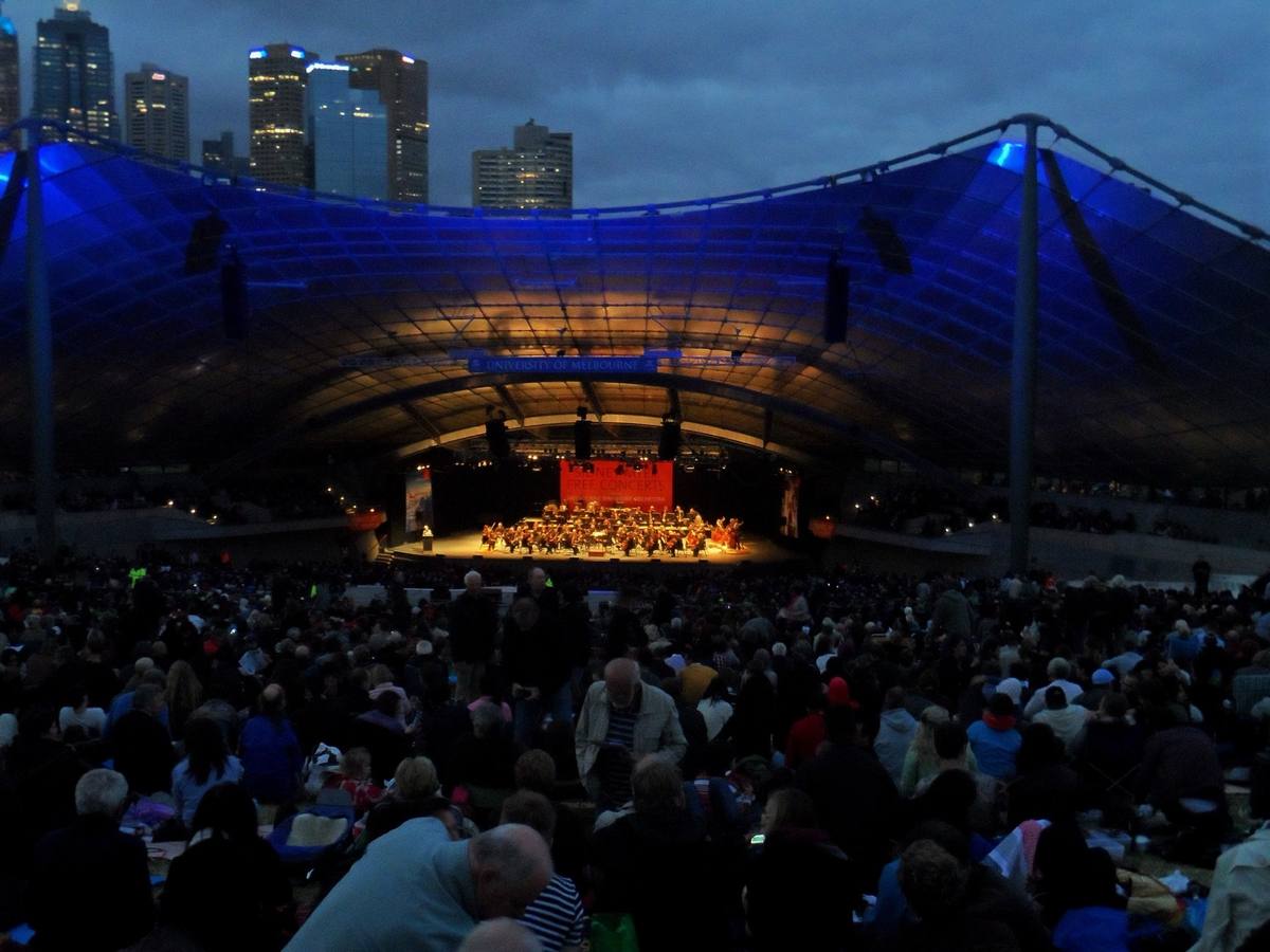A huge blue-lit canopy over an orchestra, the lawn full of people, city towers behind