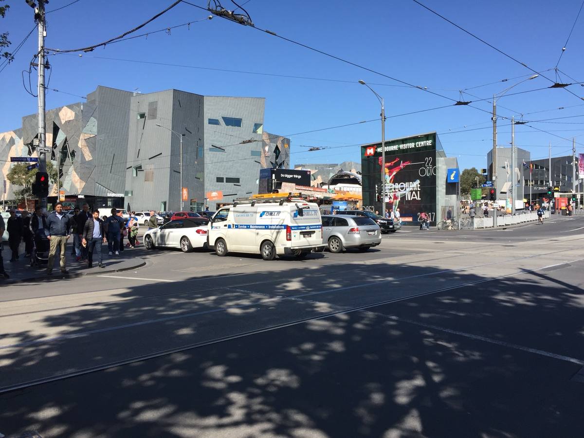 A wide city crossing with tram wires and the angular buildings of Federation Square