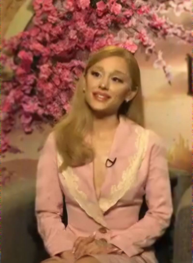 A woman with pale blonde hair in a pink outfit, smiling during an interview