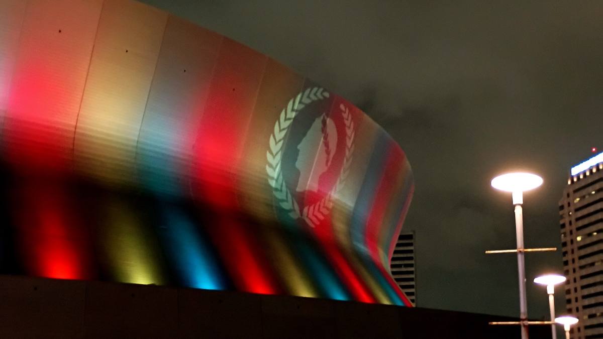 The curved side of a huge domed stadium washed in bands of red, yellow and blue light at night