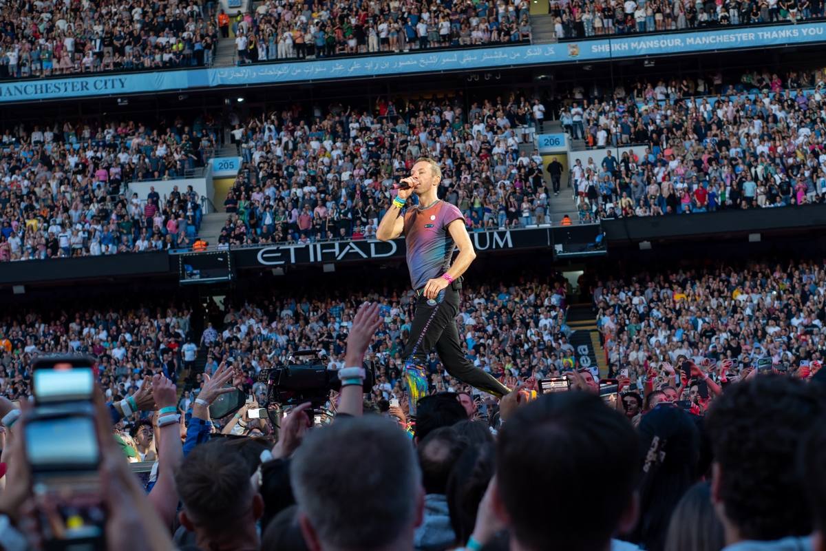 Chris Martin on a walkway in front of packed stands, the stadium name along the tier