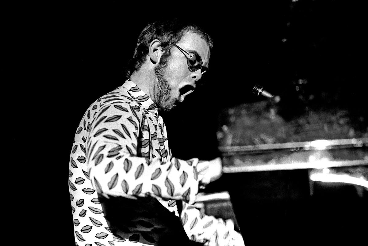 Elton John in a patterned shirt and round glasses, mouth open mid-song, playing a grand piano