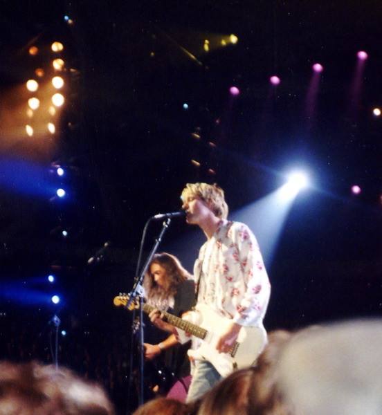 Kurt Cobain at a microphone with a white guitar under stage lights, Krist Novoselic behind him