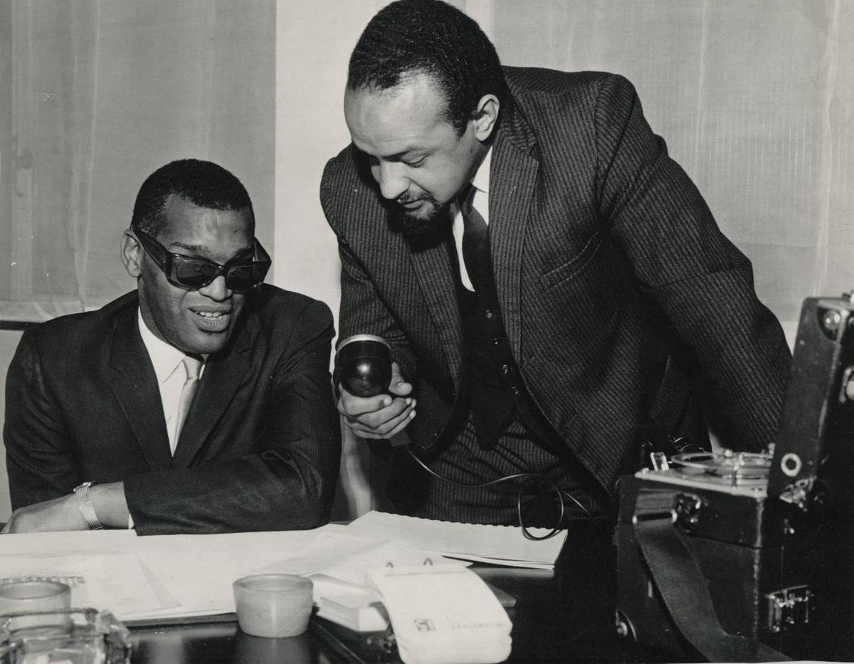 Ray Charles in dark glasses smiling at a desk while Andrew Salkey leans over him with a microphone, a tape recorder beside them