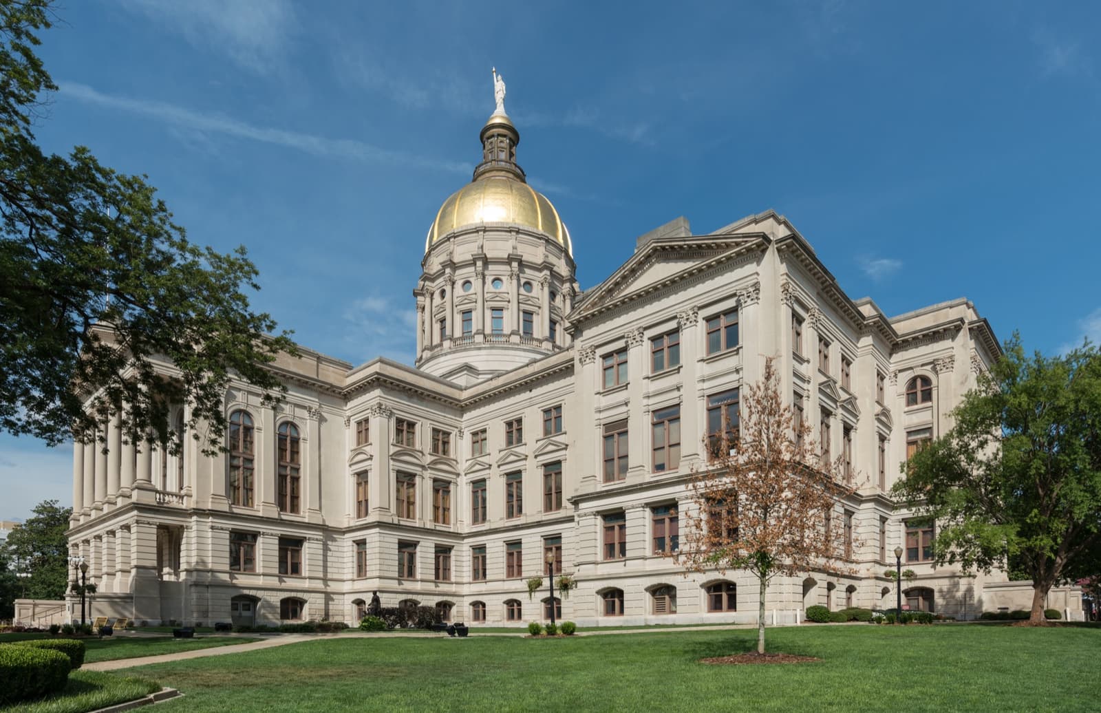 A pale stone capitol building with a gold dome under a blue sky, lawn in front