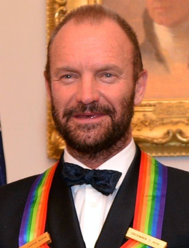 Sting in a dinner jacket and bow tie with a rainbow honours ribbon, short beard, smiling