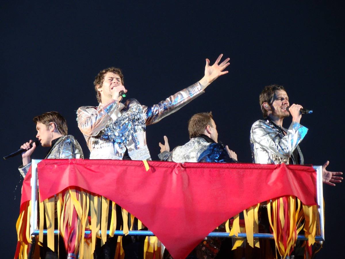 The four men in silver jackets singing behind a red-draped rail on a stadium stage at night