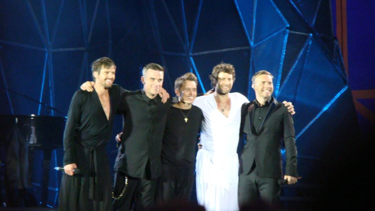 Five men in black and white standing arm in arm in front of a blue-lit faceted set