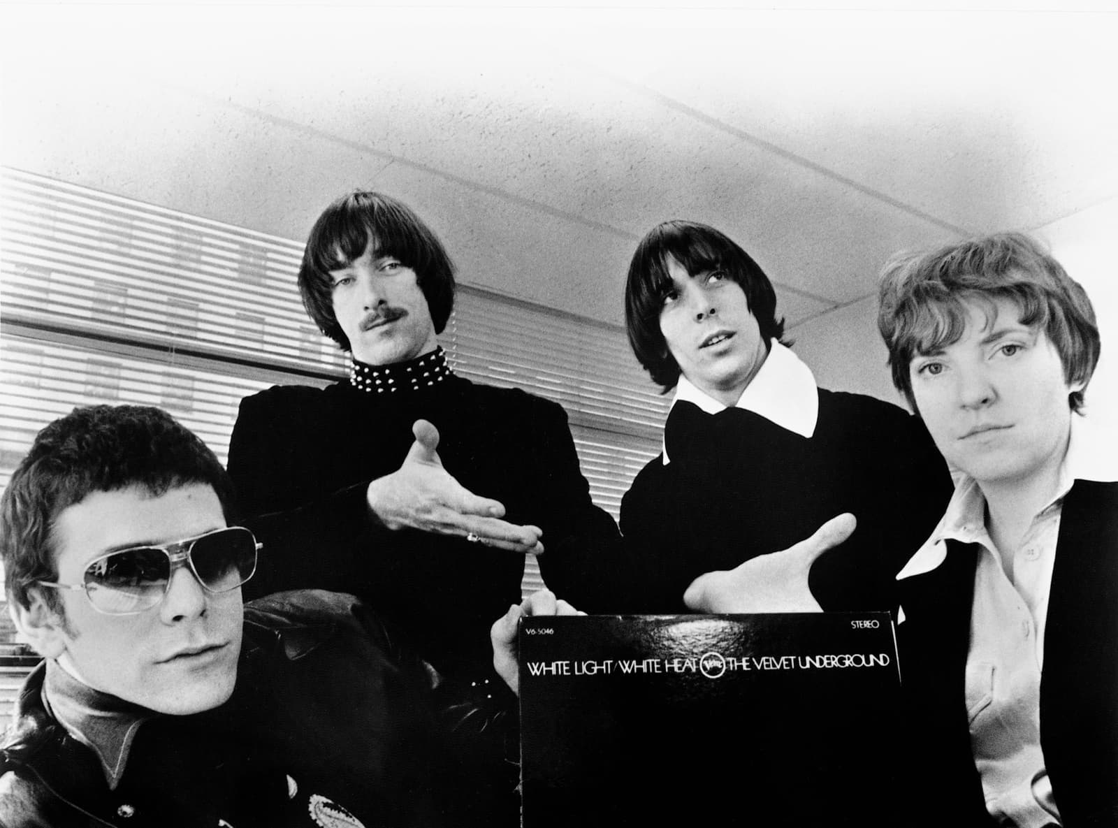Four band members in black and white, one in dark glasses in front, gesturing at an LP sleeve