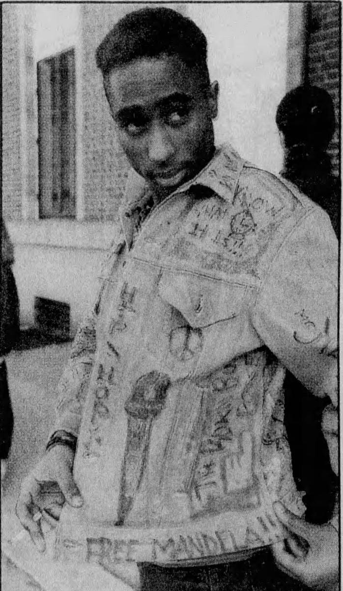 Tupac Shakur aged 17 in a denim jacket covered in hand-written words, standing outside his Baltimore school in 1988