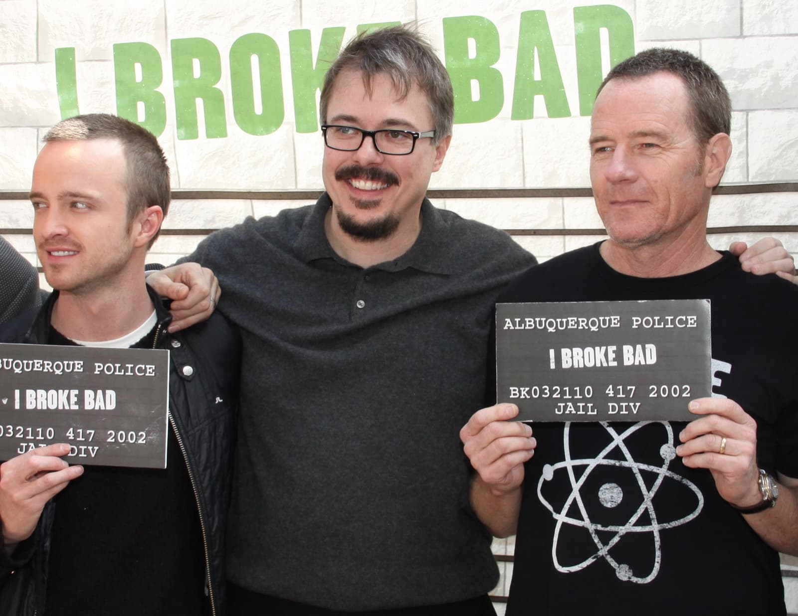 Aaron Paul, Vince Gilligan and Bryan Cranston smiling, the two actors holding mock police booking boards