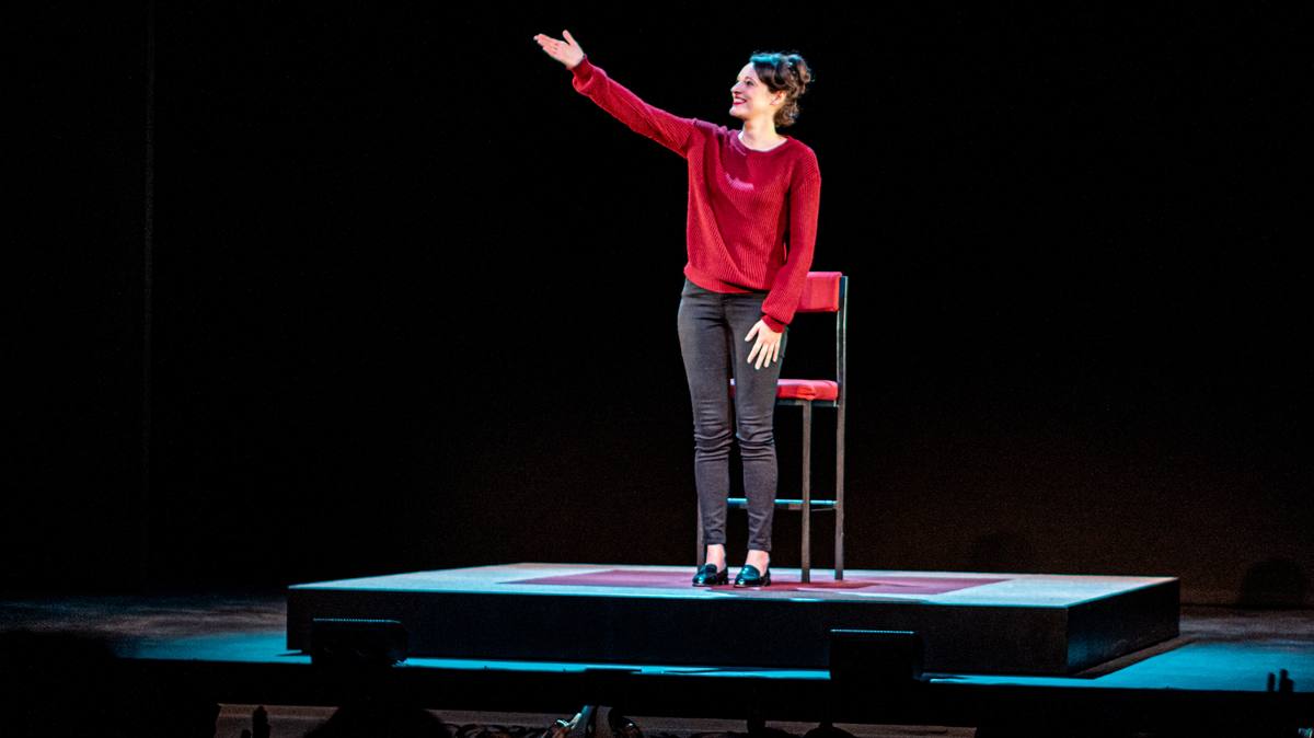 A woman in a red jumper and dark trousers stands on a small low stage beside a tall red chair, one arm raised, in a pool of light on a black stage