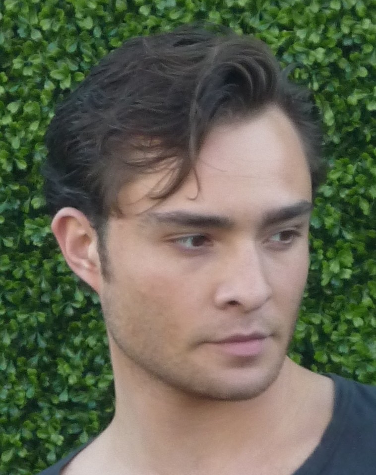 Ed Westwick, dark hair swept back, in front of a hedge