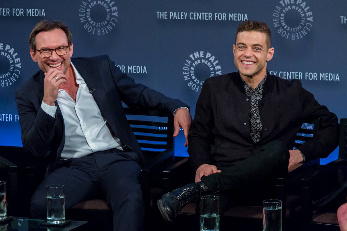 Christian Slater and Rami Malek laughing on stage, seated in armchairs