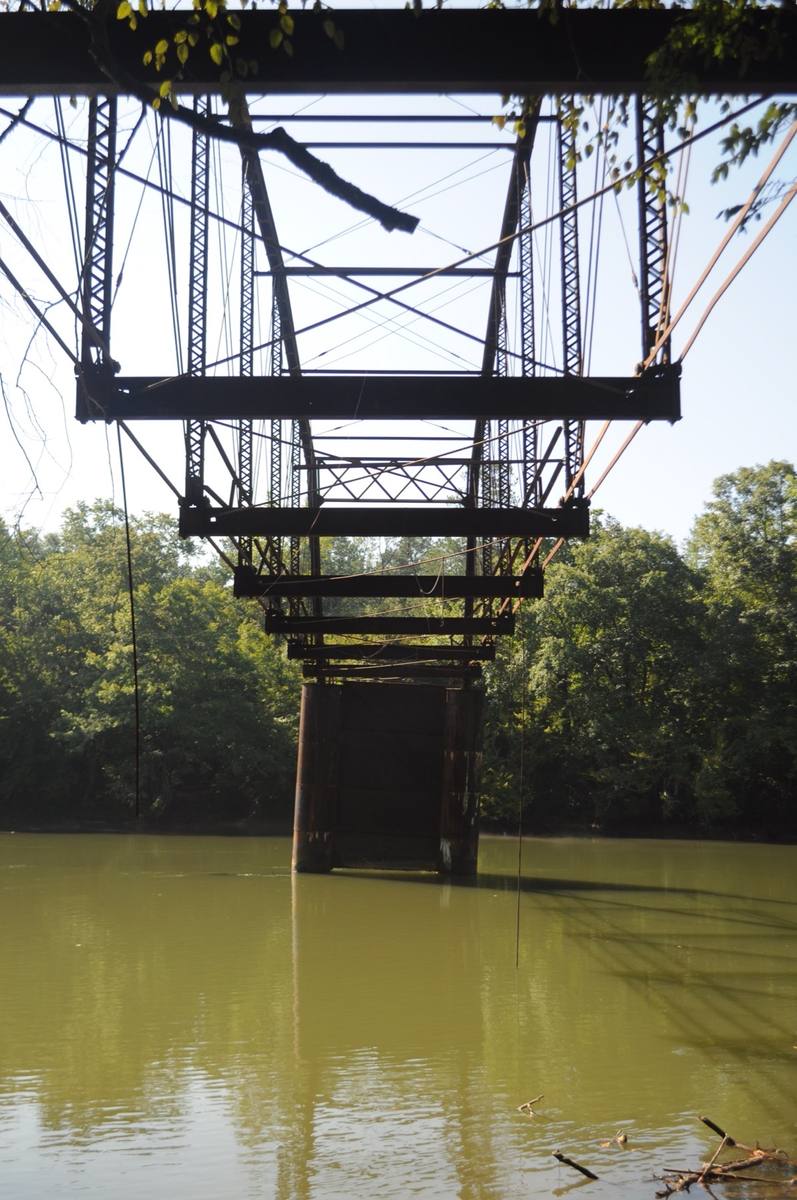 The remains of an iron truss bridge seen from below, over a green river