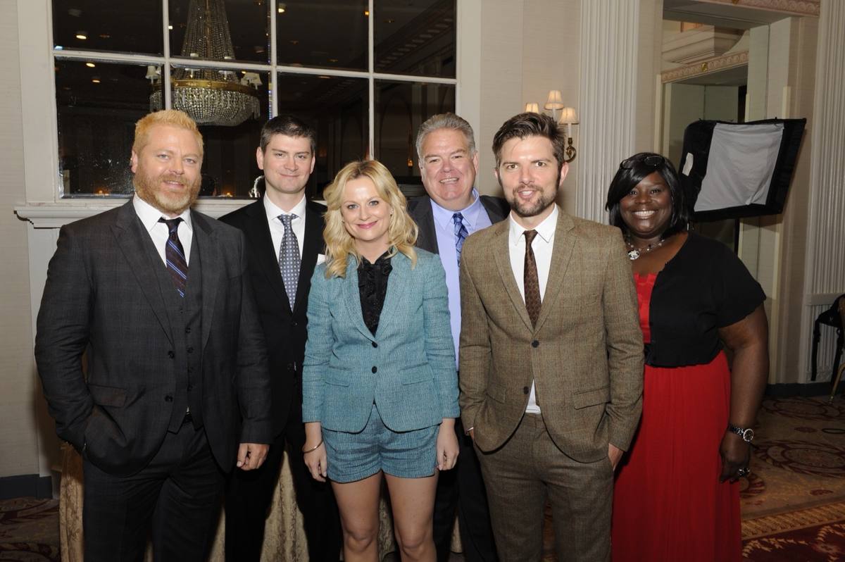 Six people in suits and dresses smiling in a hotel ballroom, a blonde woman in a pale blue suit at the centre
