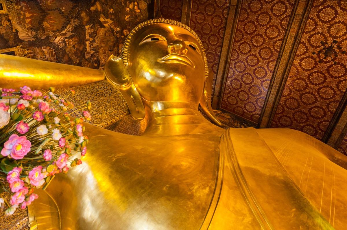 The golden face of a vast reclining Buddha against a patterned red and gold wall, pink lotus flowers in front